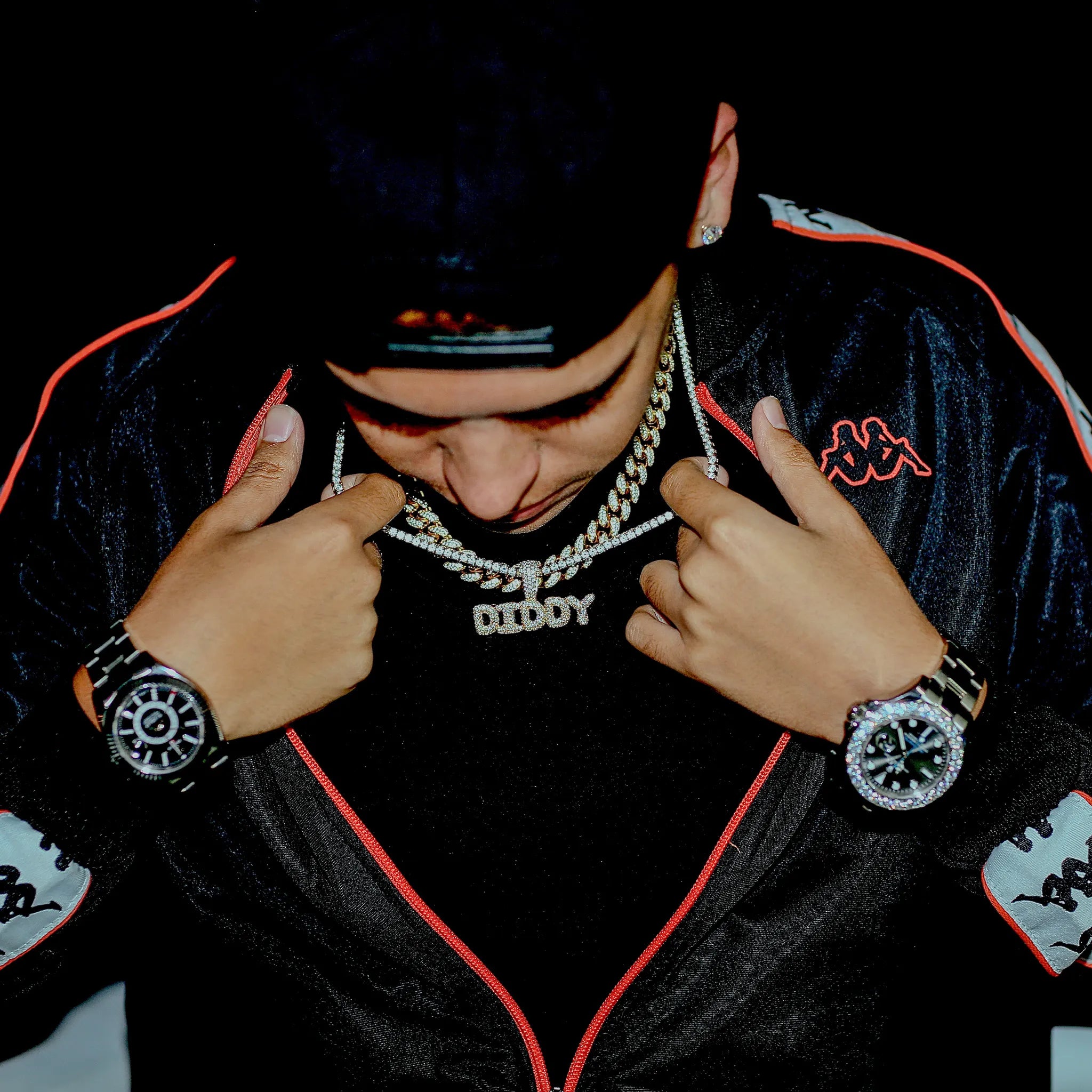 Person wearing a black jacket with red accents, a chain necklace, and watches, with a dark background.