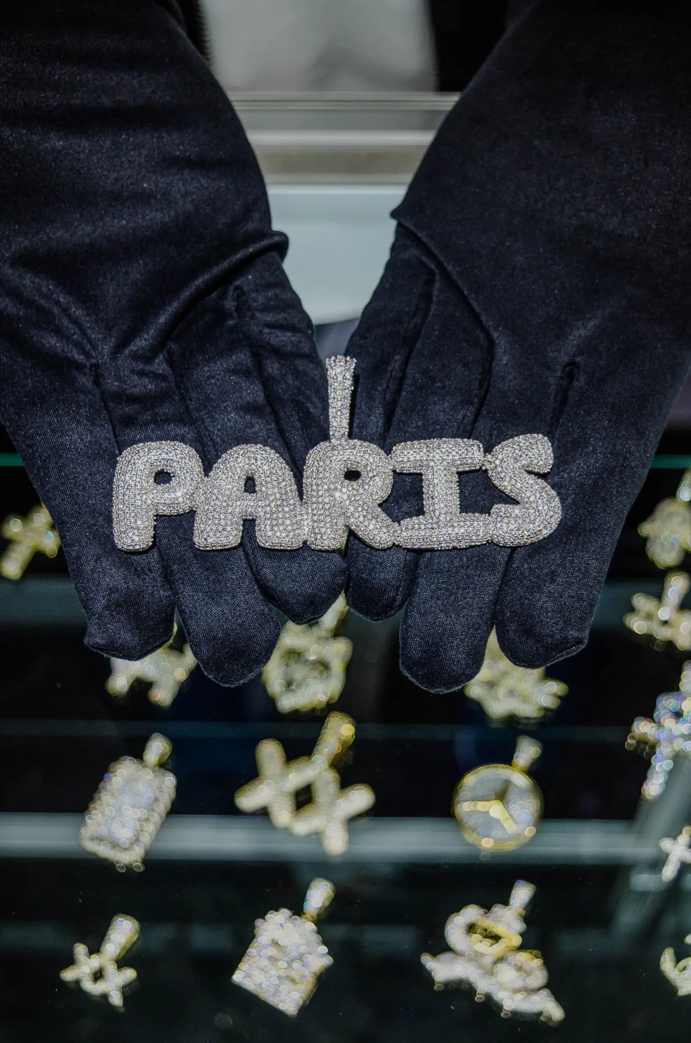 Black gloves holding a 'PARIS' sign with rhinestones against a blurred background of jewelry.