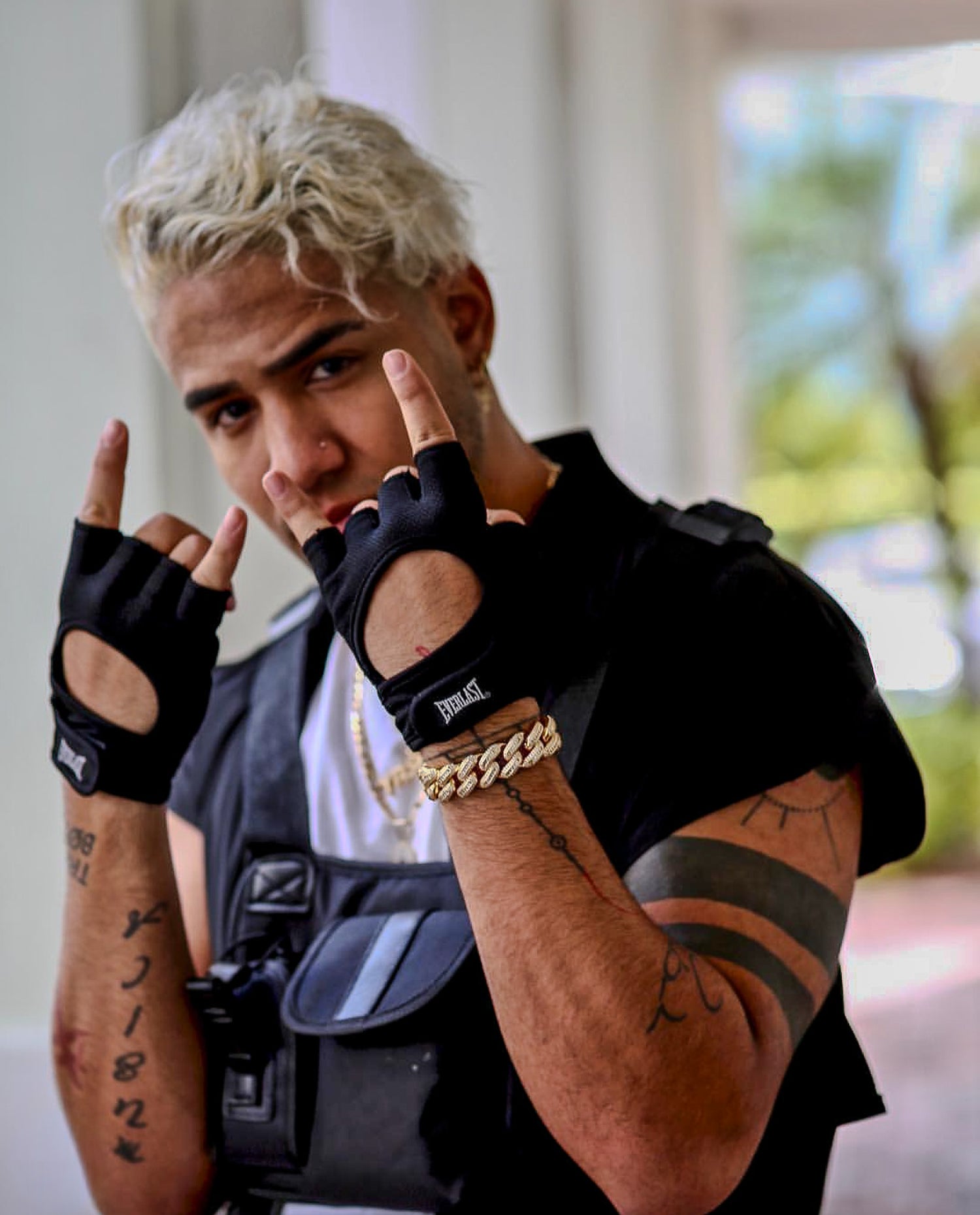 Person wearing black fingerless gloves and a black vest with tattoos on arms, making a hand gesture.
