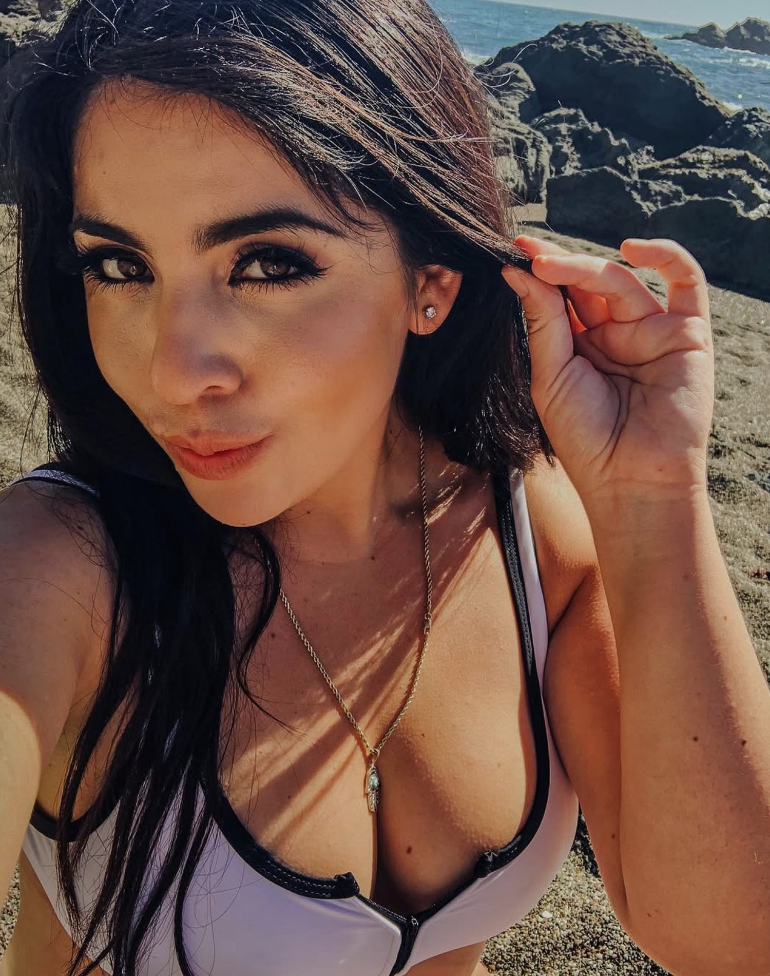 Woman with dark hair and makeup, wearing a white top, standing outdoors with rocky landscape and water in the background.