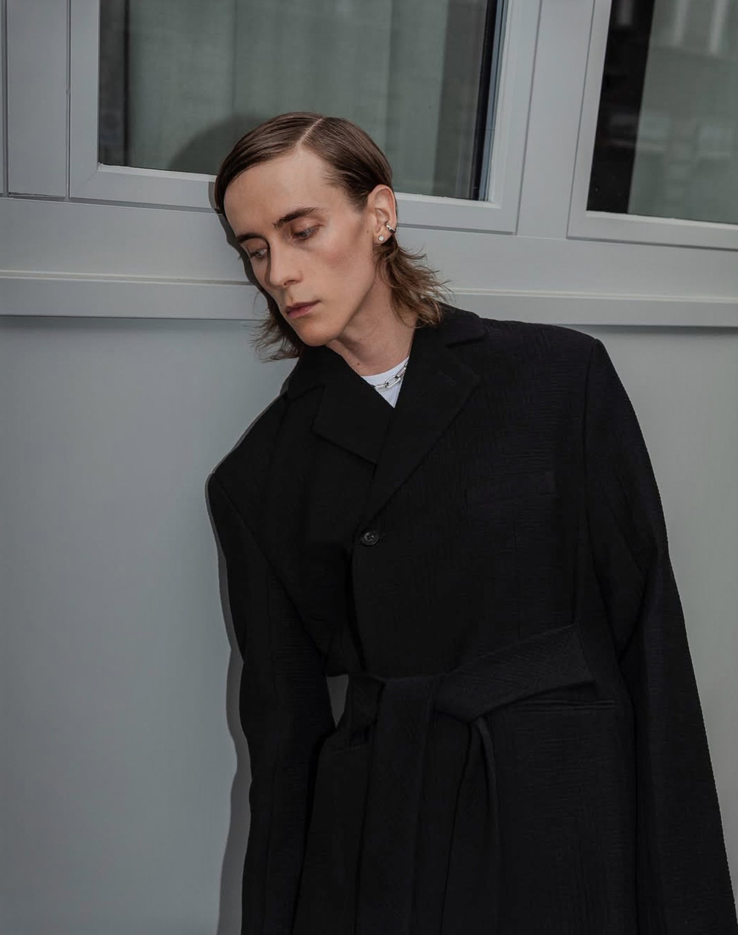 Person wearing a black coat standing against a light-colored wall with a window in the background