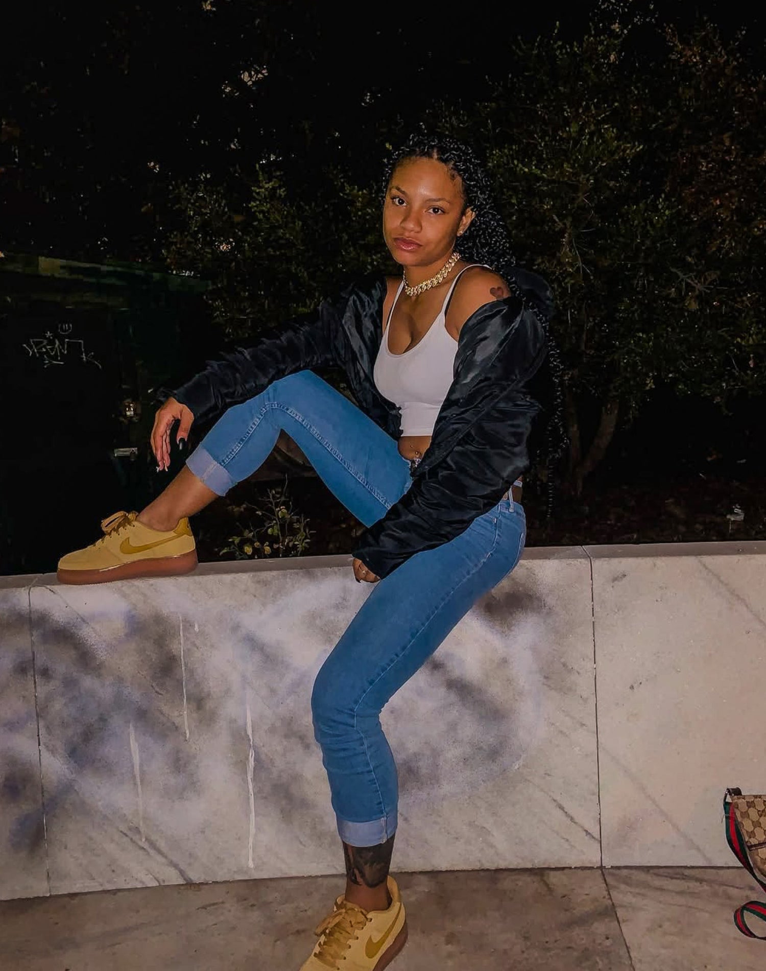 Woman sitting on a marble ledge wearing blue jeans, a black jacket, and yellow sneakers.