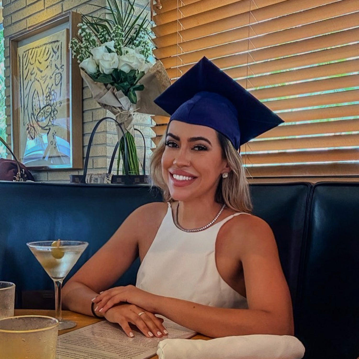 Woman in a graduation cap and gown sitting at a table with a drink, smiling.