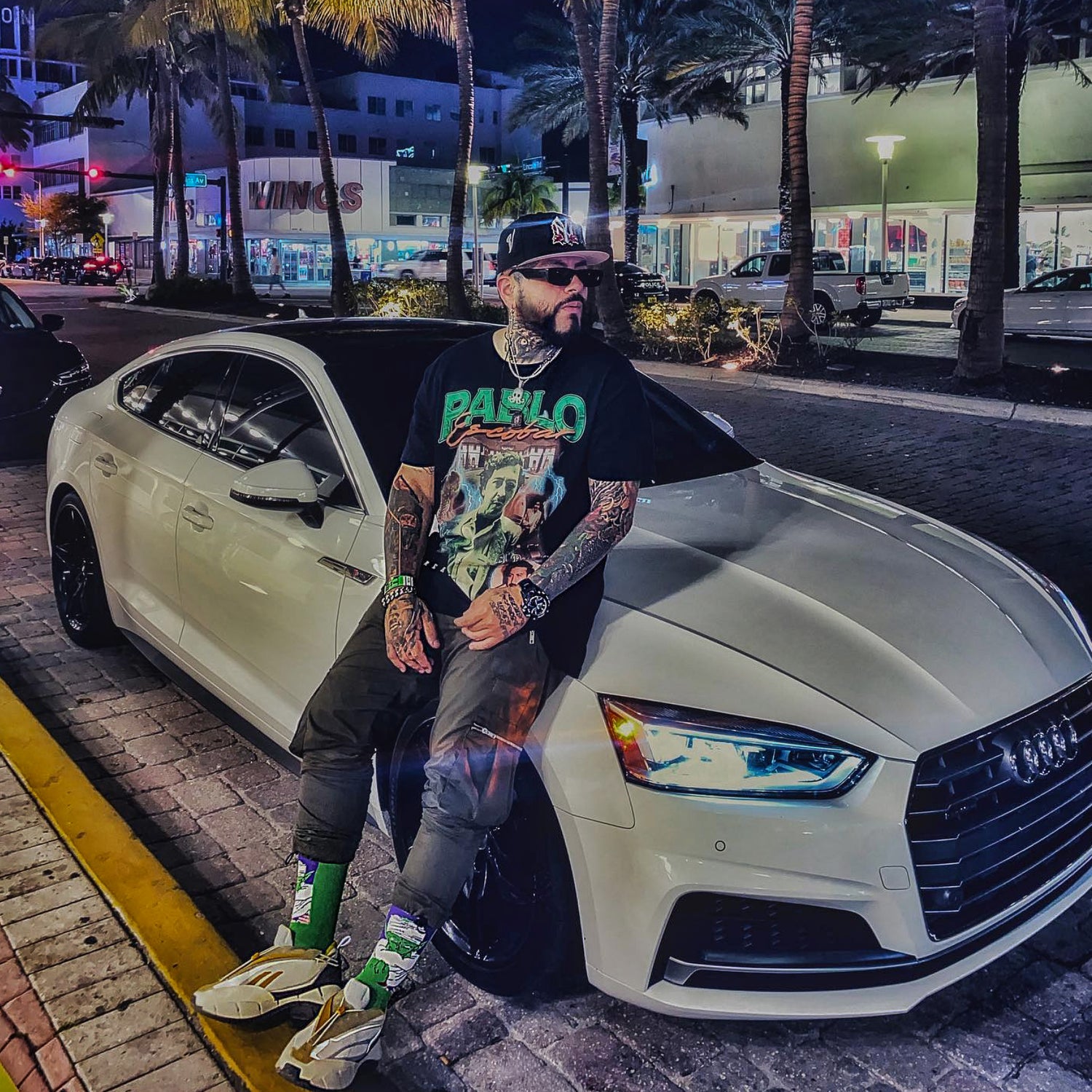 Man posing next to a white Audi car on a city street at night.