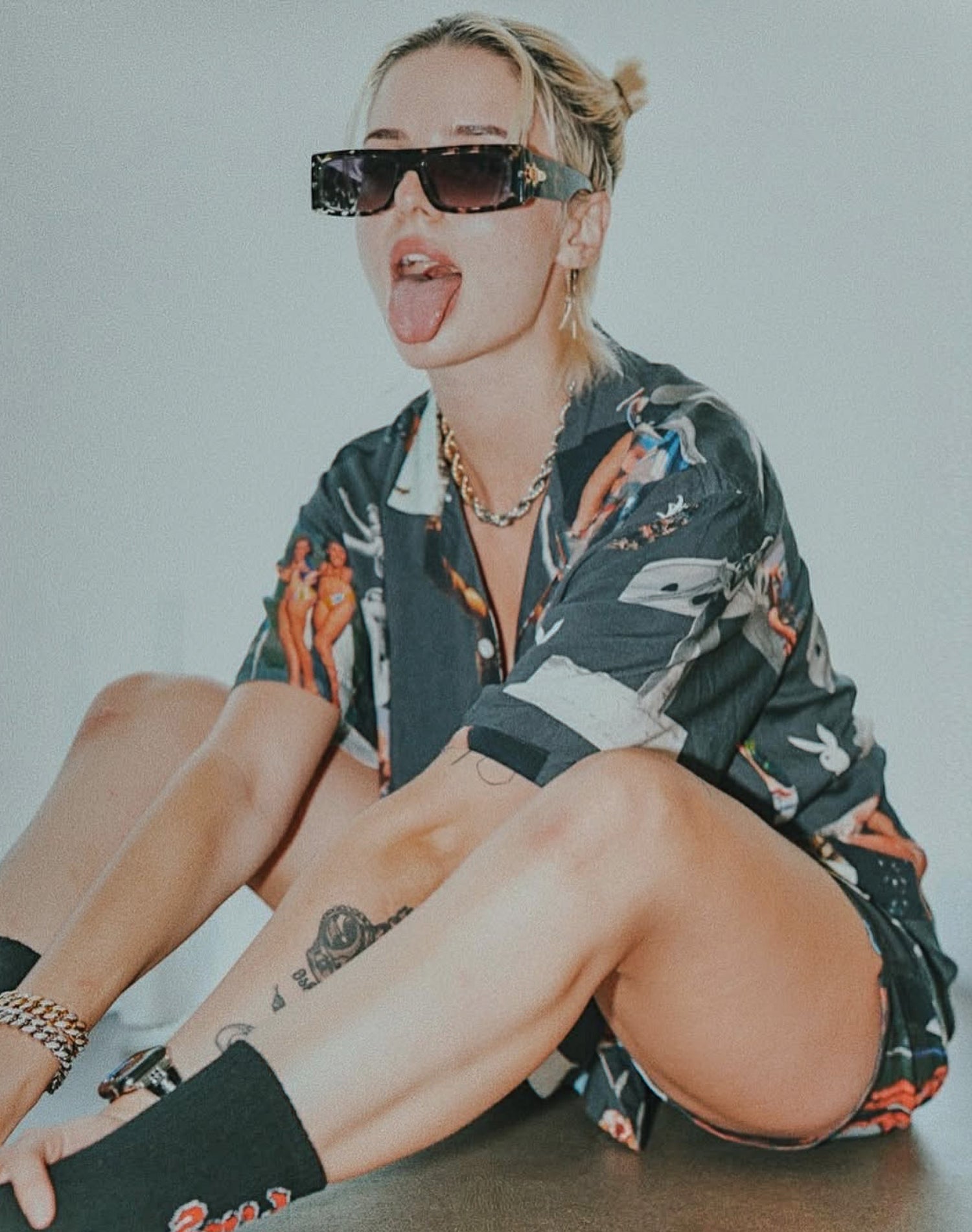 Person wearing a patterned shirt and sunglasses, sitting on the floor with a neutral background