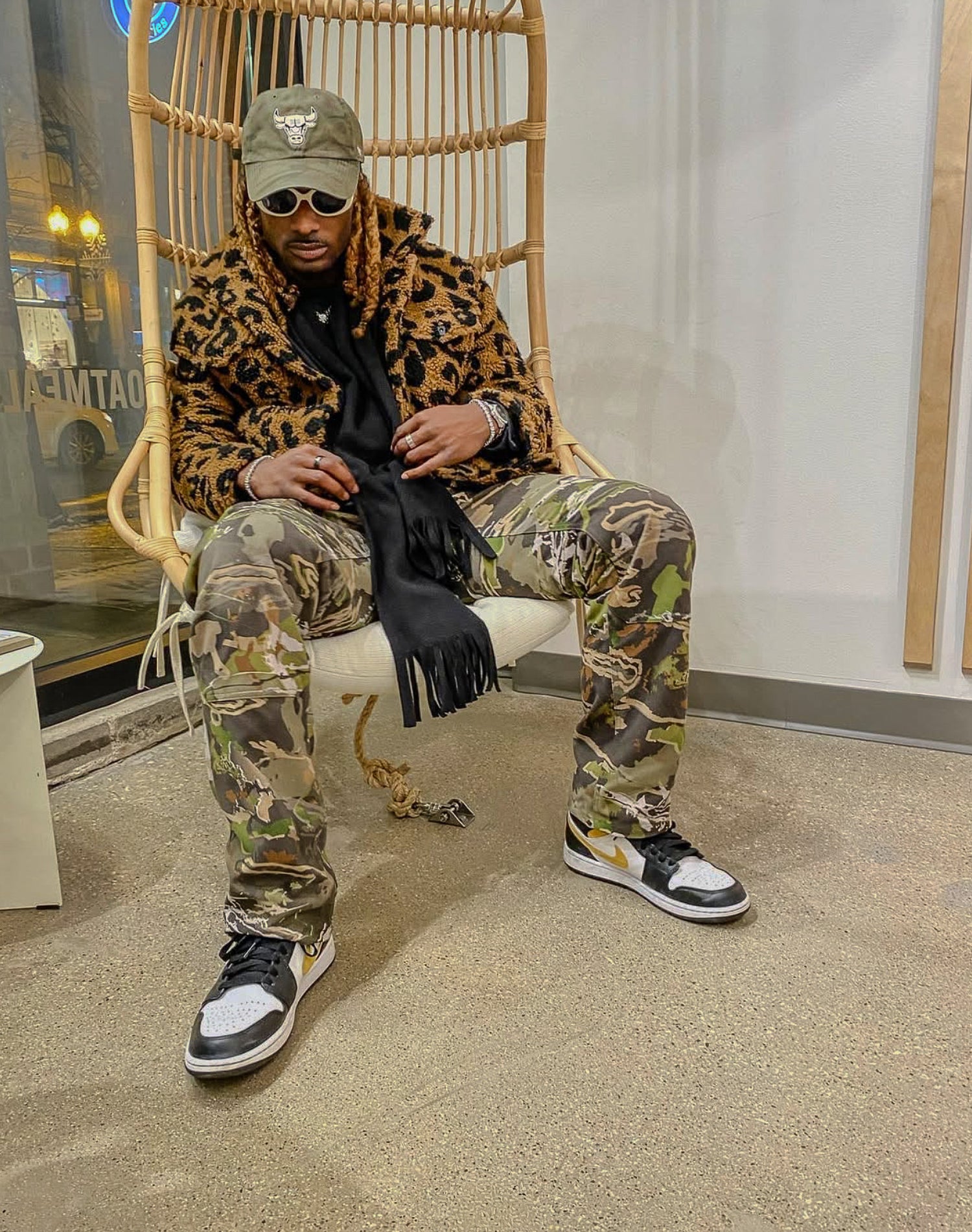 Person wearing a leopard print jacket, camouflage pants, and black sneakers sitting on a wicker chair.